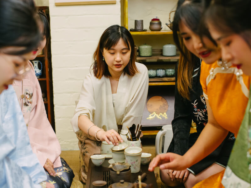 Several women sit around a table at a tea tasting ceremony, with tea equipment in front of them